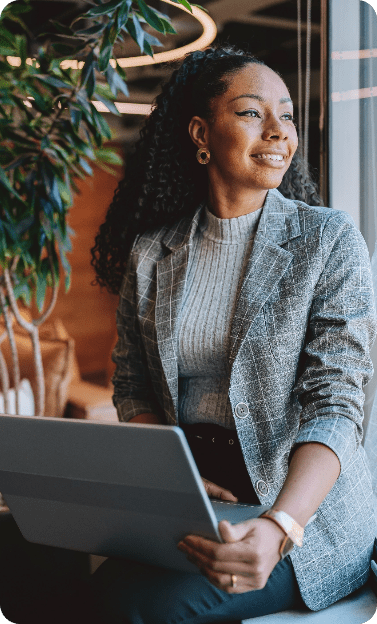 A smiling Black woman with curly hair, wearing a gray plaid blazer and gold hoop earrings, sits by a window holding an open laptop on her lap and gazes outside.