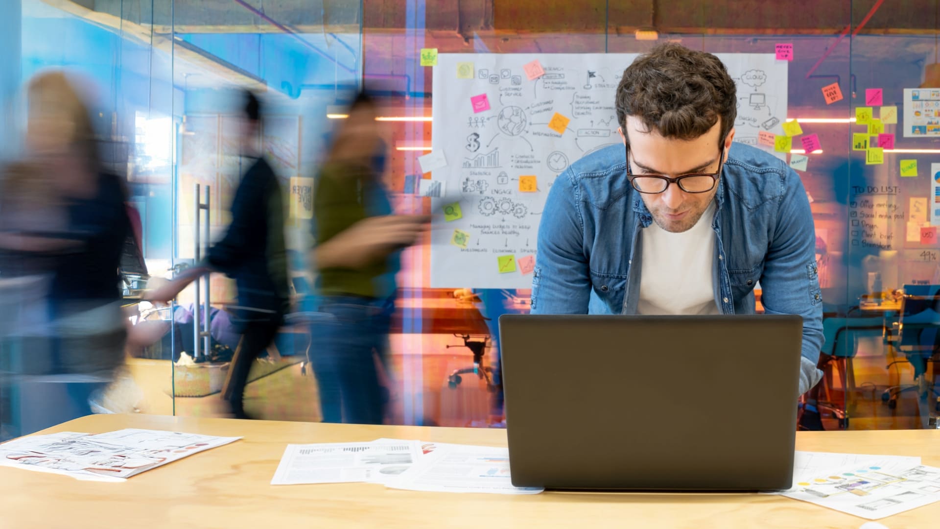 Man working on laptop in busy office with notes on glass wall